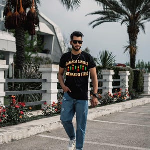 May include: A black short-sleeved t-shirt featuring a graphic of colourful candles and the text "JAPANESE CANDLES BEWARE OF FIRE!" paired with blue denim jeans and white trainers. The shirt has a crew neckline and a central printed design.