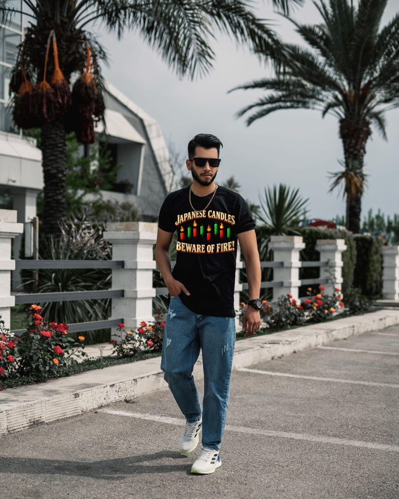 May include: A black short-sleeved t-shirt featuring a graphic of colourful candles and the text "JAPANESE CANDLES BEWARE OF FIRE!" paired with blue denim jeans and white trainers. The shirt has a crew neckline and a central printed design.