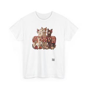 May include: A white t-shirt featuring a graphic print of three alpacas in white, tan, and dark brown. Each alpaca wears a colorful patterned blanket and floral headpiece. The alpacas stand amidst a cluster of flowers at the base of the design.