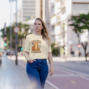 May include: A cream-colored short-sleeve t-shirt featuring a graphic of an orange cat sitting in a lotus position. The text above the cat reads LESS DRAMA and the text below reads MORE KARMA. The shirt is paired with blue denim jeans.