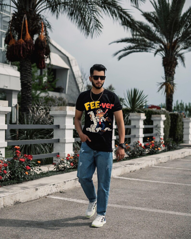 May include: A black short-sleeved t-shirt featuring a graphic of a cartoon character holding a long receipt with the text FEE-ver in yellow and red letters, paired with light blue distressed denim jeans and white athletic trainers.