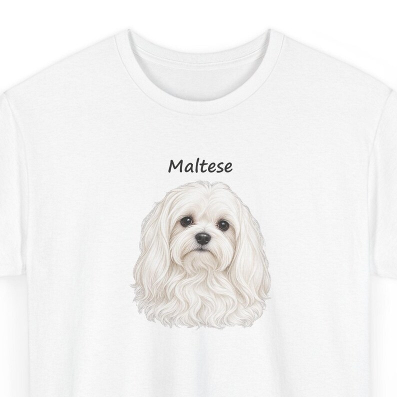 May include: A white t-shirt featuring a detailed illustration of a fluffy white Maltese dog centered on the chest. Above the dog's head, the word "Maltese" is printed in a simple, black, sans-serif font. The shirt has a crew neckline.