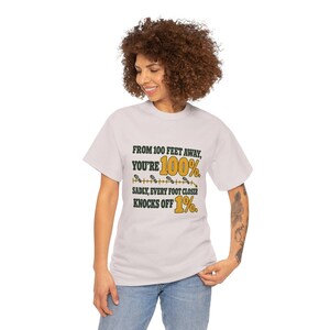 May include: A light gray short-sleeve t-shirt featuring dark green and yellow text that reads: FROM 100 FEET AWAY, YOU'RE 100%. SADLY, EVERY FOOT CLOSER KNOCKS OFF 1%. The design includes a yellow line with small footprint icons.