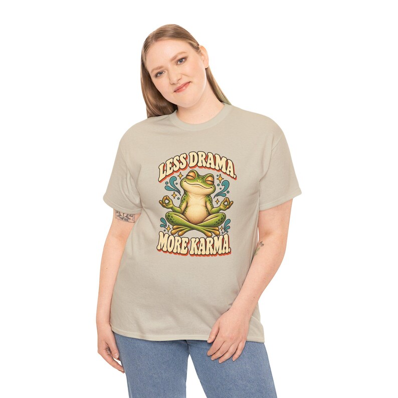 May include: A beige short-sleeve t-shirt featuring a graphic of a green frog sitting in a lotus position with eyes closed. The text above the frog reads LESS DRAMA and the text below reads MORE KARMA in a stylized, rounded font.