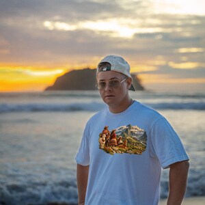 May include: A white short-sleeved t-shirt featuring a colorful graphic print of people in traditional clothing with llamas in a mountainous landscape. The shirt is worn with a light-colored baseball cap and round-framed glasses.