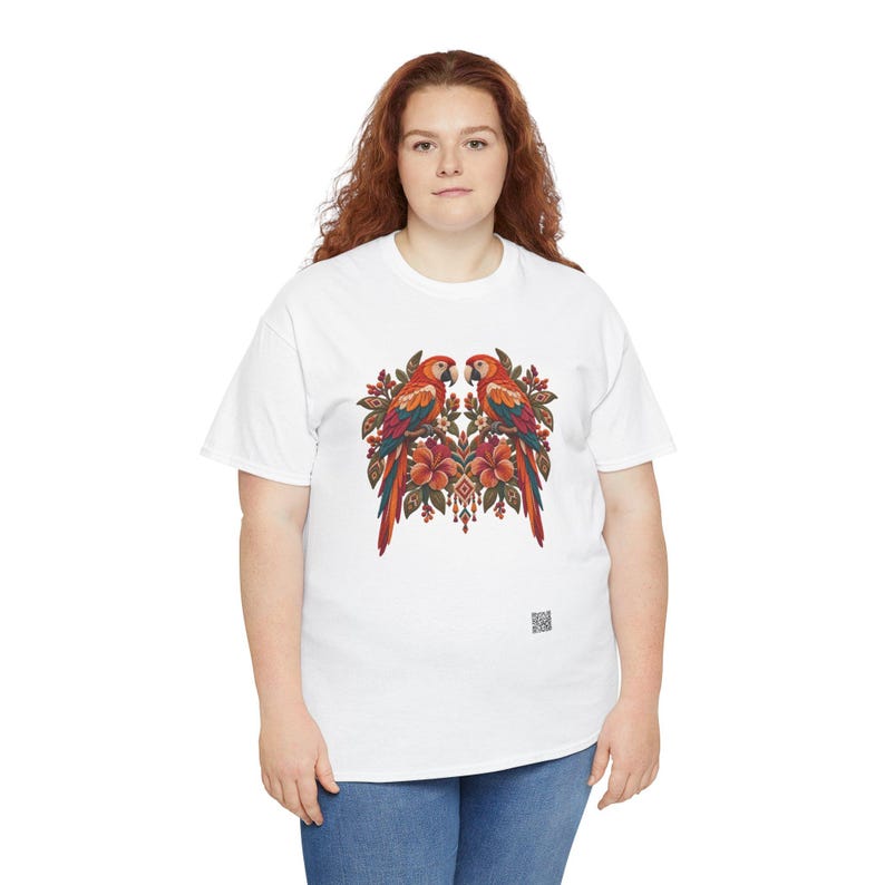 May include: A white short-sleeved t-shirt featuring a central graphic of two symmetrical parrots perched among tropical flowers and foliage in shades of red, orange, and green. A small square QR code is printed near the bottom right hem.
