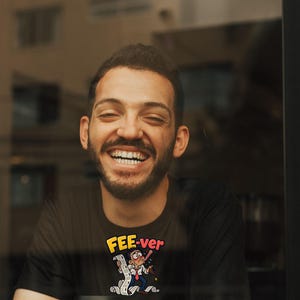 May include: A black short-sleeved t-shirt featuring a graphic print of a cartoon character with a thermometer in their mouth, holding a long receipt, with the yellow and red text "FEE-ver" printed above the illustration.