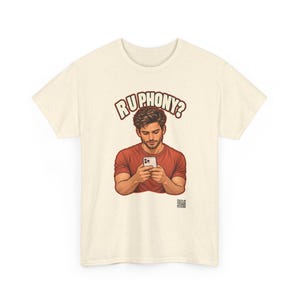 May include: A cream-colored short-sleeve t-shirt featuring a graphic of a person in a red shirt looking at a smartphone. Above the figure, the text reads "RU PHONY?" in bold letters. A small QR code is printed near the bottom right of the graphic.
