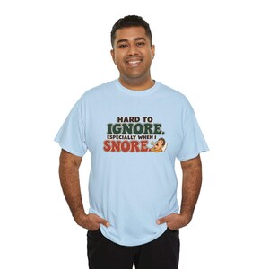 May include: A light blue short-sleeve t-shirt featuring the text "HARD TO IGNORE. ESPECIALLY WHEN I SNORE." in dark brown and green lettering, accompanied by a small graphic of a person sleeping and snoring on a white pillow.