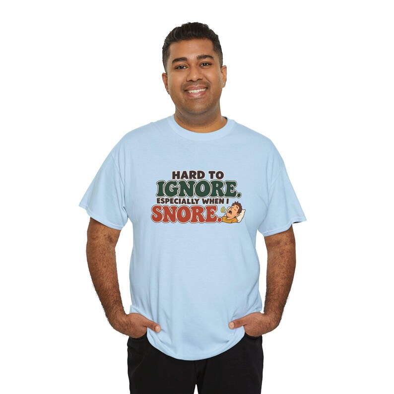 May include: A light blue short-sleeve t-shirt featuring the text "HARD TO IGNORE. ESPECIALLY WHEN I SNORE." in dark brown and green lettering, accompanied by a small graphic of a person sleeping and snoring on a white pillow.