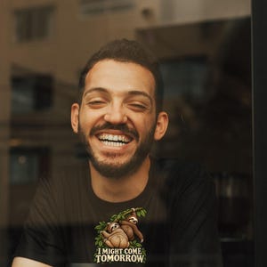 Pode incluir: Uma t-shirt preta de manga curta com um gr&aacute;fico de duas pregui&ccedil;as penduradas num ramo verde com folhas. Abaixo das pregui&ccedil;as, o texto "I MIGHT COME TOMORROW" aparece em letras mai&uacute;sculas brancas sobre o tecido preto da pe&ccedil;a.