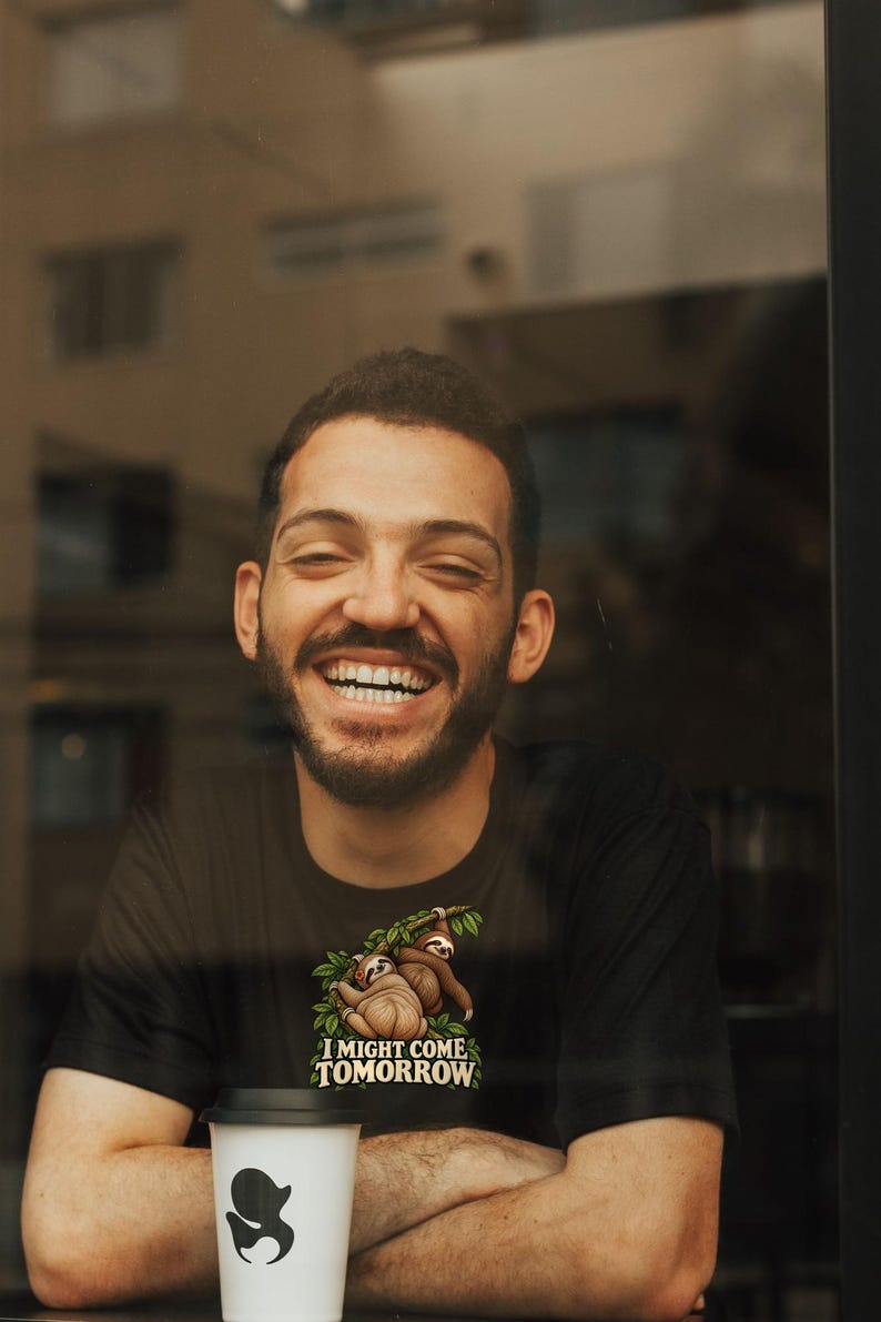 Pode incluir: Uma t-shirt preta de manga curta com um gr&aacute;fico de duas pregui&ccedil;as penduradas num ramo verde com folhas. Abaixo das pregui&ccedil;as, o texto "I MIGHT COME TOMORROW" aparece em letras mai&uacute;sculas brancas sobre o tecido preto da pe&ccedil;a.