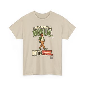 Op de afbeelding: Een beige t-shirt met korte mouwen en een afbeelding van een lopend persoon. De tekst luidt I WAS BORN TO WALK. NOT TO WORK. De persoon draagt een groen overhemd, een wit t-shirt en een bruine broek. Een kleine QR-code staat rechtsonder.