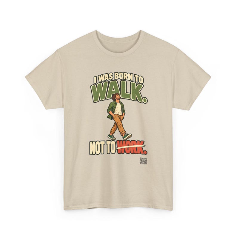Op de afbeelding: Een beige t-shirt met korte mouwen en een afbeelding van een lopend persoon. De tekst luidt I WAS BORN TO WALK. NOT TO WORK. De persoon draagt een groen overhemd, een wit t-shirt en een bruine broek. Een kleine QR-code staat rechtsonder.