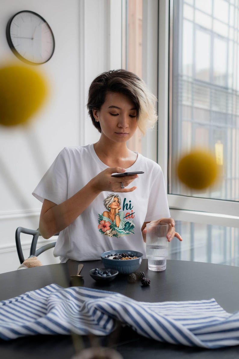 Puede incluir: Una camiseta blanca de manga corta con un estampado gr&aacute;fico de una persona con el pelo corto, una flor tropical y olas del mar, con el texto "hi" en una fuente estilizada. La camiseta tiene un cuello redondo.