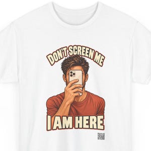 Op de afbeelding: Een wit t-shirt met een afbeelding van een persoon die een smartphone voor het gezicht houdt. De tekst erboven luidt DON'T SCREEN ME en de tekst eronder luidt I AM HERE in vetgedrukte letters. Een kleine QR-code staat rechtsonder.