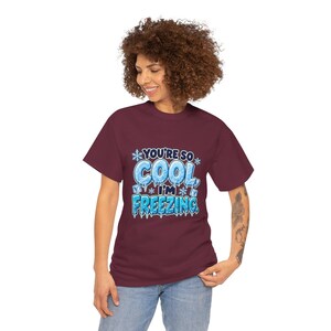 May include: A maroon short-sleeve t-shirt featuring a graphic print with the text "YOU'RE SO COOL, I'M FREEZING." The design includes light blue icy lettering, white snowflakes, and small ice cubes, creating a winter-themed visual.