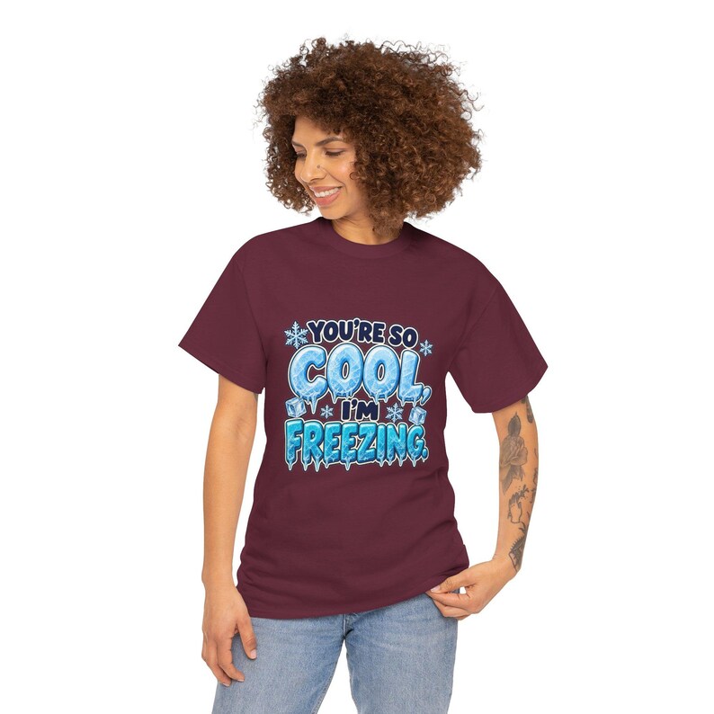 May include: A maroon short-sleeve t-shirt featuring a graphic print with the text "YOU'RE SO COOL, I'M FREEZING." The design includes light blue icy lettering, white snowflakes, and small ice cubes, creating a winter-themed visual.