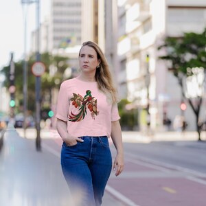 May include: A light pink short-sleeved t-shirt featuring a detailed graphic of a quetzal bird perched on a branch with cocoa pods. The bird has vibrant green, red, and white plumage with a long, flowing tail. The design includes small geometric patterns.