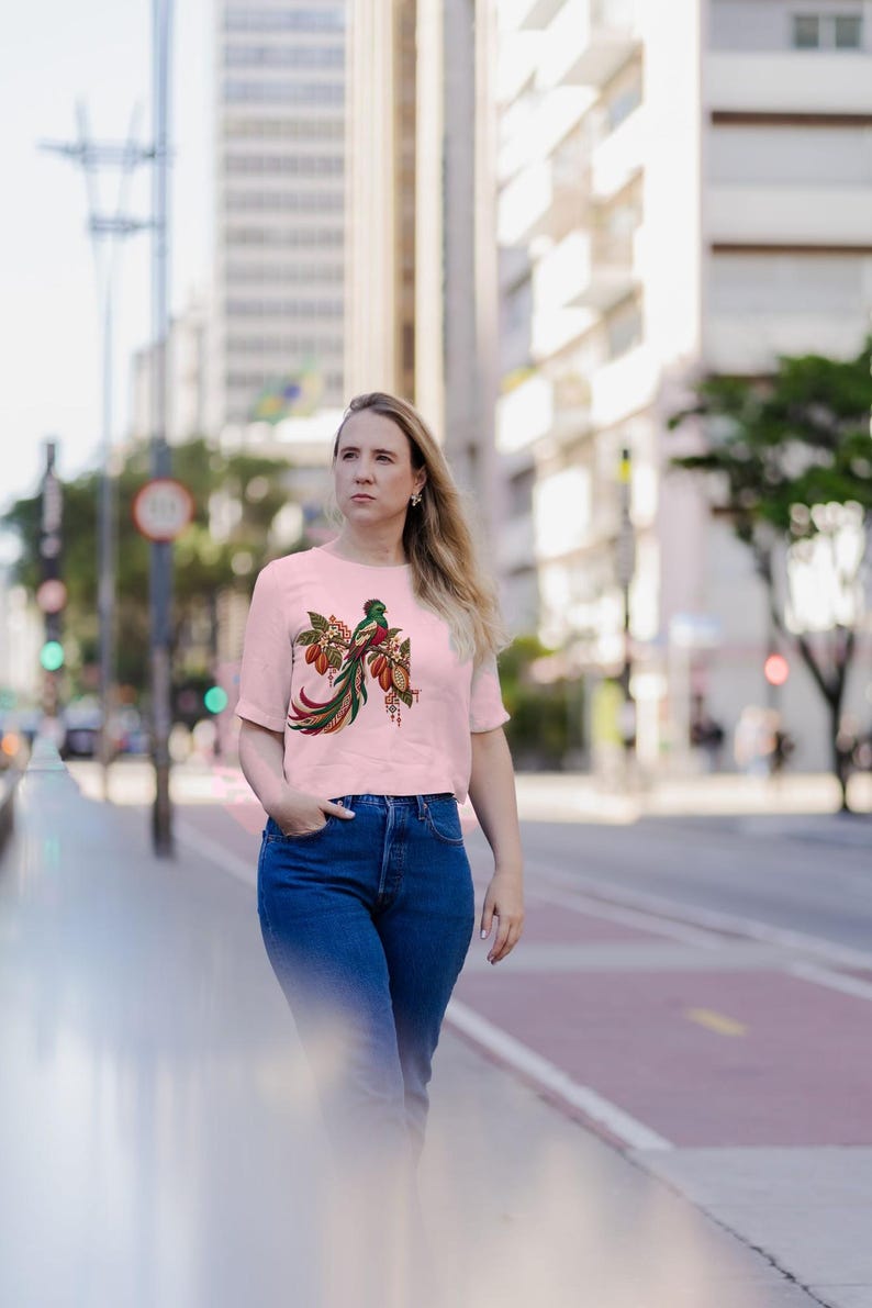 May include: A light pink short-sleeved t-shirt featuring a detailed graphic of a quetzal bird perched on a branch with cocoa pods. The bird has vibrant green, red, and white plumage with a long, flowing tail. The design includes small geometric patterns.