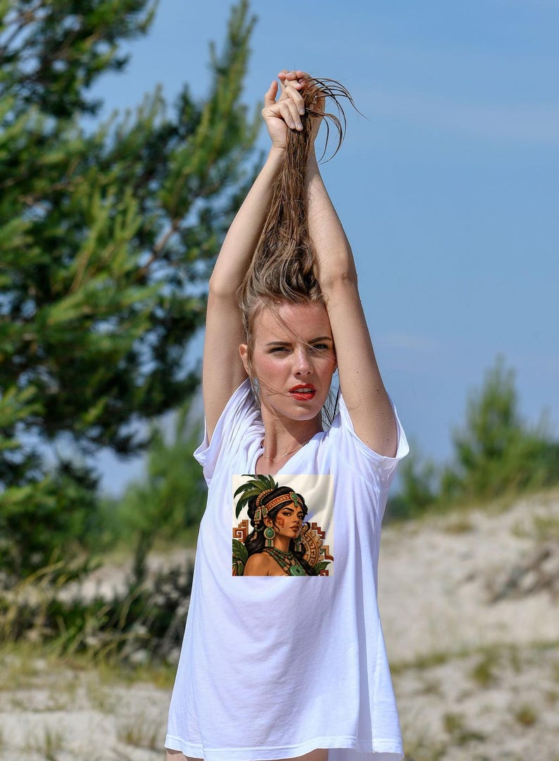 Puede incluir: Una camiseta blanca de manga corta con un estampado gr&aacute;fico cuadrado en el pecho. El dise&ntilde;o muestra a una persona de cabello oscuro con un tocado adornado con plumas verdes, grandes pendientes circulares y un collar sobre un fondo color crema.