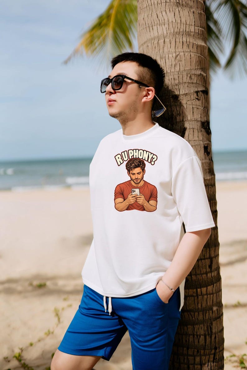 May include: A white short-sleeved t-shirt featuring a graphic print of a person in a rust-colored shirt looking at a smartphone, with the text "RUPHONY?" arched above the illustration in a bold, light-colored font with a dark outline.