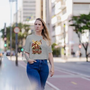 May include: A light gray short-sleeved t-shirt featuring a graphic of a golden angel blowing a trumpet, surrounded by red ribbons and green holly leaves, with the text "JOY TO THE WORLD" printed in a decorative red and gold font.