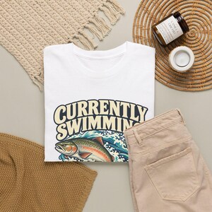 May include: A white t-shirt featuring the text CURRENTLY SWIMMING in large cream-colored letters above a graphic illustration of a colorful trout fish jumping out of blue and white ocean waves with detailed water splashes.