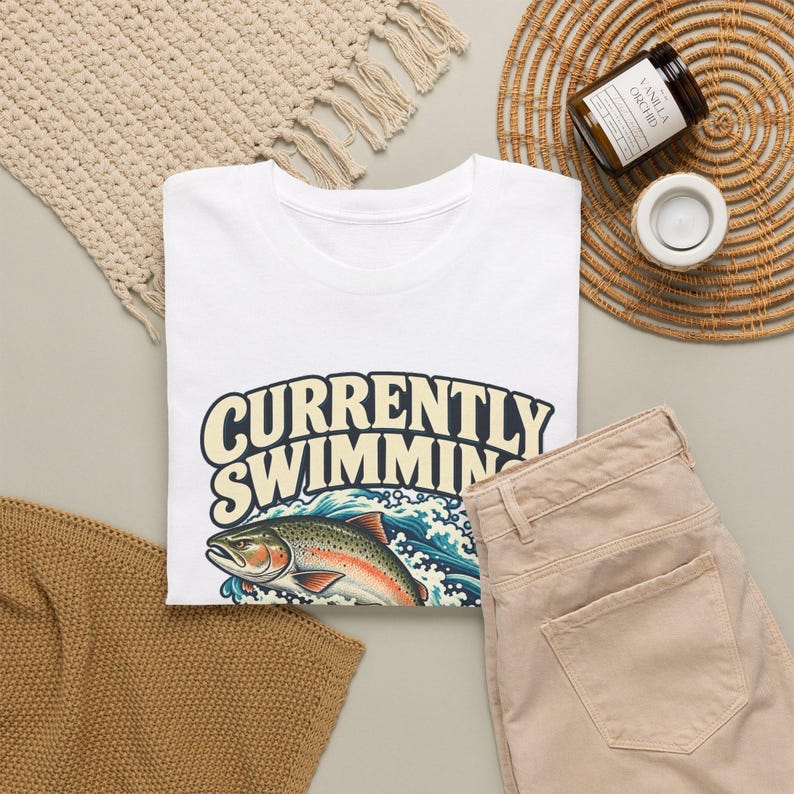 May include: A white t-shirt featuring the text CURRENTLY SWIMMING in large cream-colored letters above a graphic illustration of a colorful trout fish jumping out of blue and white ocean waves with detailed water splashes.