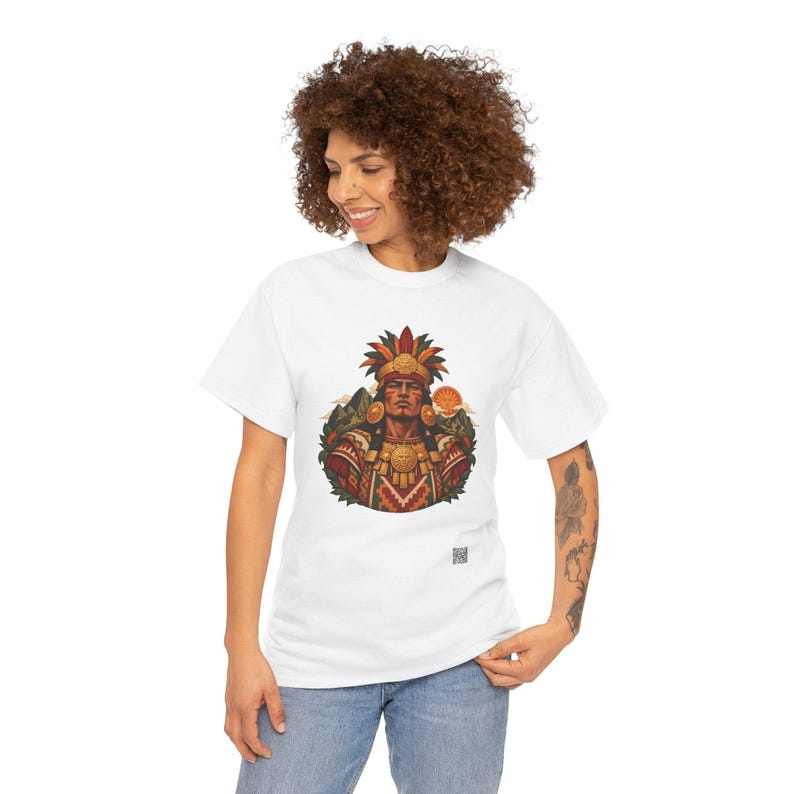 May include: A white t-shirt featuring a central graphic of a person wearing a colorful feathered headdress and ornate traditional clothing. The design includes earth tones, red, and yellow accents, with a small square QR code printed below the image.
