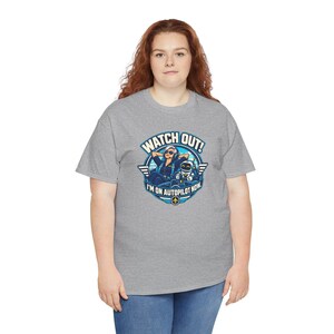 May include: A heather gray short-sleeve t-shirt featuring a graphic of a pilot relaxing in a cockpit with a robot co-pilot. The text reads WATCH OUT! I'M ON AUTOPILOT NOW. The design includes blue, white, and yellow accents.