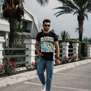 Puede incluir: Una camiseta negra de manga corta con un gr&aacute;fico de un pez saltando sobre una presa con el texto NO DAMNED MAN-MADE DAM WILL STOP ME!, combinada con pantalones vaqueros azul claro desgastados y zapatillas deportivas blancas con rayas negras.