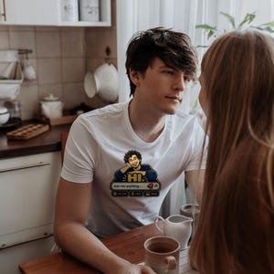 May include: A white short-sleeved t-shirt featuring a graphic print of a person with curly hair resting their head on a hand. Below the figure is a chat interface box with the text "HI" and "Ask me anything..." along with icons.