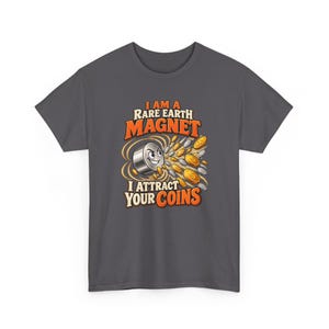 May include: A dark gray t-shirt featuring a graphic of a cartoon magnet with a face, surrounded by gold and silver coins. The text reads, I AM A RARE EARTH MAGNET I ATTRACT YOUR COINS, in orange and white lettering with a bold, stylized font.