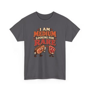 May include: A dark gray t-shirt featuring a graphic of two anthropomorphic steak pieces. One steak holds a magnifying glass, looking at the other. The text reads I AM MEDIUM LOOKING FOR RARE in orange and red lettering with a small red heart.