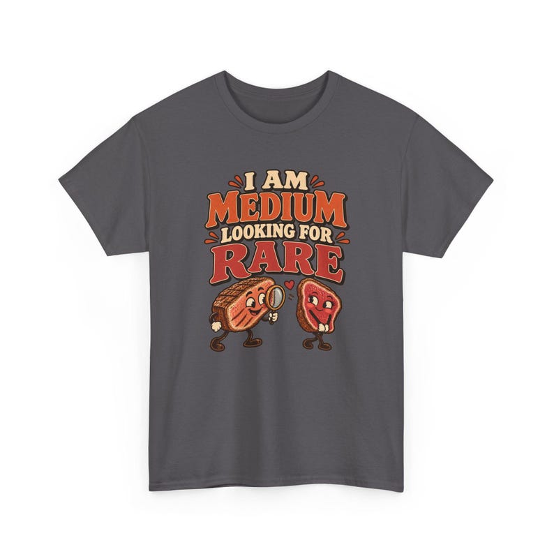 May include: A dark gray t-shirt featuring a graphic of two anthropomorphic steak pieces. One steak holds a magnifying glass, looking at the other. The text reads I AM MEDIUM LOOKING FOR RARE in orange and red lettering with a small red heart.