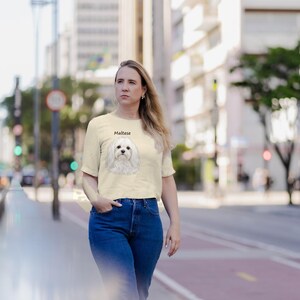 May include: A cream-colored short-sleeve t-shirt featuring a graphic illustration of a white Maltese dog centered on the chest with the word Maltese printed in black text above the dog's head. The shirt is paired with blue denim jeans.