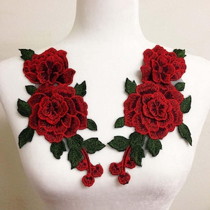 Puede incluir: Un par de parches bordados florales simétricos que presentan rosas de color rojo intenso con pétalos en capas y hojas de color verde oscuro. El intrincado trabajo de aguja muestra sombreados detallados en las flores y el follaje, creando una apariencia texturizada sobre una base de malla transparente.