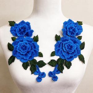 Op de afbeelding: Een paar symmetrische geborduurde bloemapplicaties met levendige blauwe, gelaagde roosachtige bloemen en donkergroene bladeren en stengels. Het gedetailleerde borduurwerk toont verfijnde texturen van de bloemblaadjes en het blad in twee spiegelbeelden.