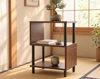 May include: A two-tier wooden side table with a dark walnut finish and black cylindrical metal supports. The top shelf holds a small bonsai tree in a ceramic pot, while the lower shelf contains a closed book, an incense holder, and a ceramic cup.