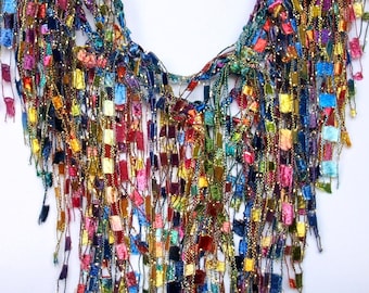 May include: A colorful crocheted necklace with a fringe of multi-colored yarn. The necklace is made of small squares of yarn in various shades of blue, green, red, yellow, and gold.