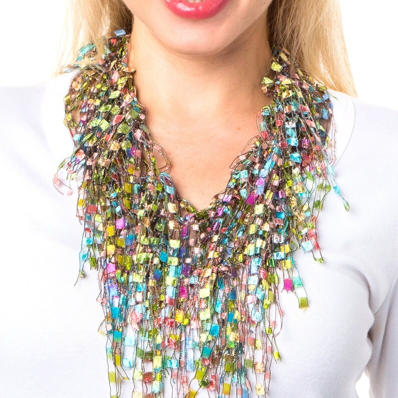 May include: A colorful, multi-layered, fringe necklace made of yarn in shades of green, blue, pink, and purple. The necklace is worn by a person with a white shirt.