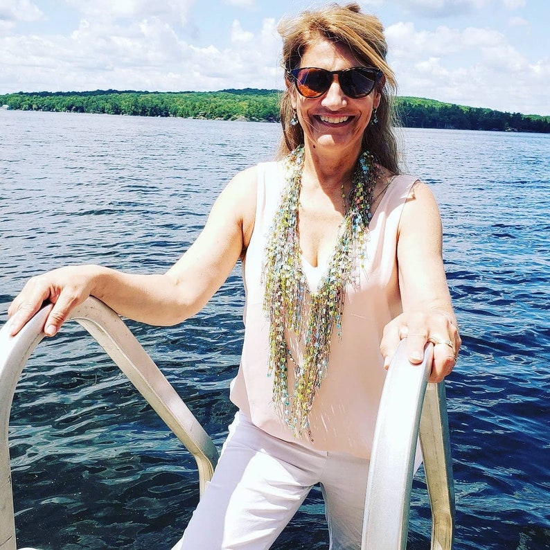 May include: A woman wearing sunglasses and a pink tank top with a long, beaded necklace. She is standing on a dock over a body of water.