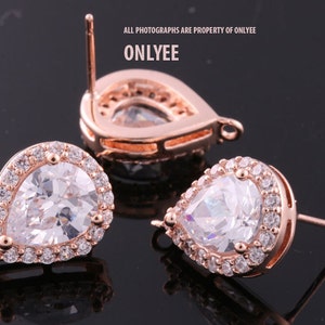 May include: A pair of rose gold teardrop earrings with clear cubic zirconia stones. The earrings have a halo setting and a post back.
