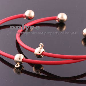May include: Two red silicone bracelets with gold-toned metal balls and a charm. The bracelets are layered on top of each other.