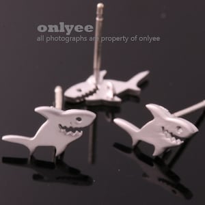 May include: A pair of silver shark stud earrings. The sharks are facing forward with their mouths open, showing their teeth. The earrings are on a black background.