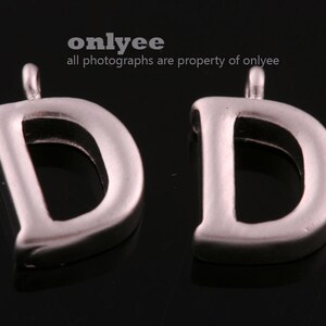 Könnte beinhalten: Zwei silberne Buchstaben-Anhänger in Form des Buchstabens D mit einer Öse oben zum Anbringen an eine Halskette oder ein Armband.