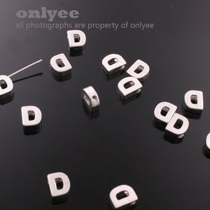 Könnte beinhalten: Silberne Buchstaben-Perlen in Form des Buchstabens D mit einem Loch in der Mitte. Die Perlen sind auf einer schwarzen Oberfläche verstreut.