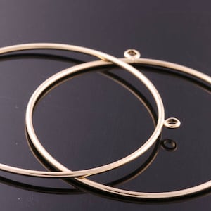 May include: Two gold-colored metal hoop earrings with small loops at the top for attaching to ear wires. The hoops are round and have a shiny finish. The earrings are displayed on a dark surface.