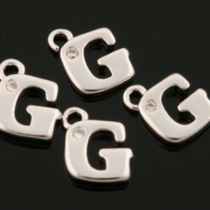 Puede incluir: Cuatro colgantes de plata con forma de letra G, cada uno con un strass transparente en el centro.